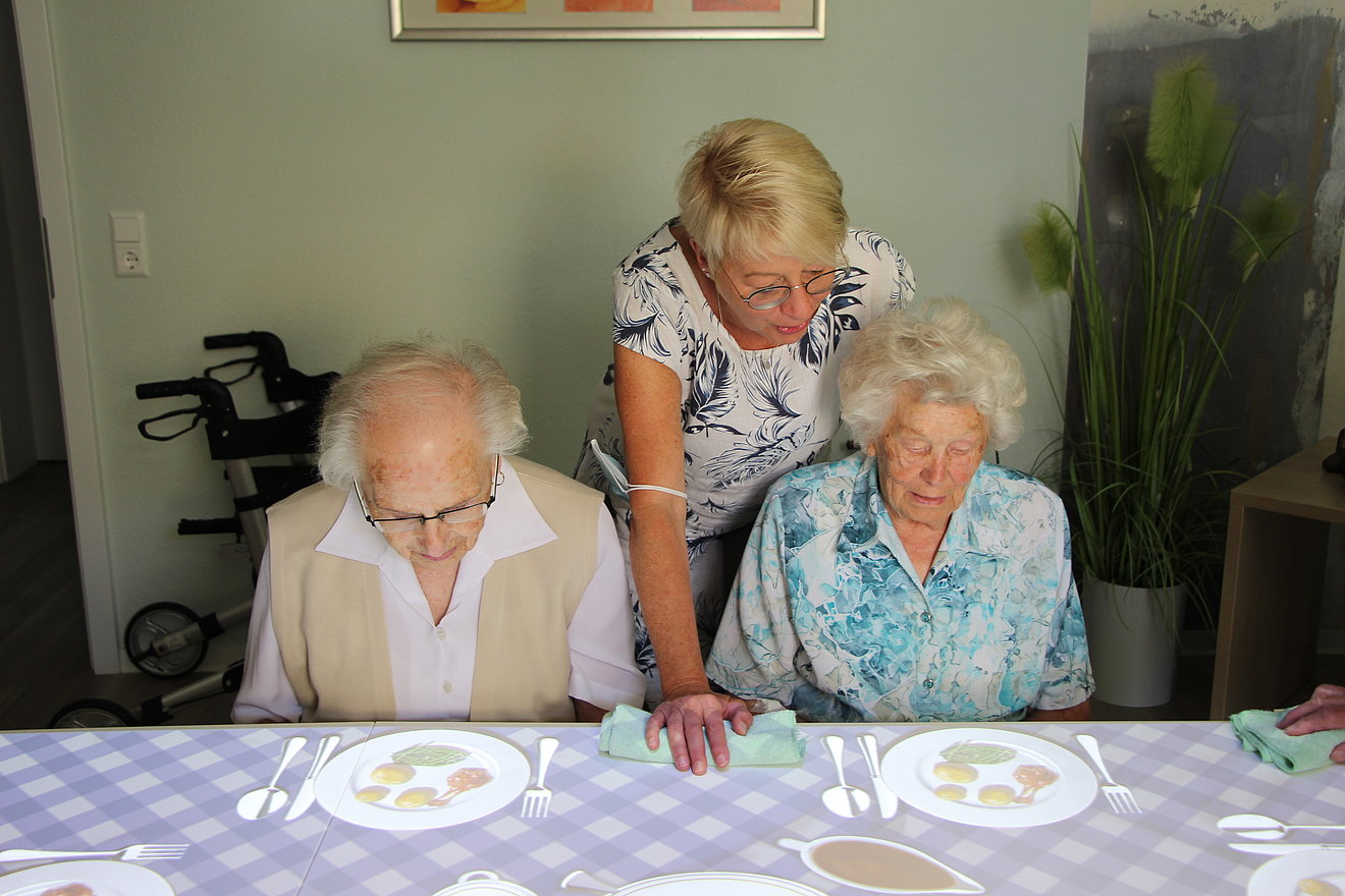 Zwei Seniorinnen sitzen an einem Tisch, auf den Bilder von Tellern mit Essen projiziert werden. eine Frau steht hinter ihnen und deutet auf die Bilder.