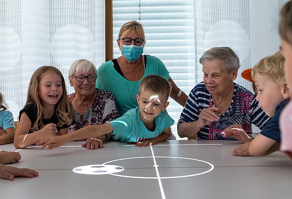 Zwei Seniorinnen, eine Frau mit Mund-Nasen-Schutz und drei Kinder beugen sich über einen Tisch, auf den ein Fußballspiel projiziert wird.