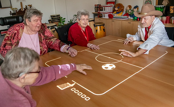 Drei Frauen und ein Mann sitzen um einen eckigen Tisch auf den ein Fußballfeld projiziert wird. Sie beugen sich über das Spielfeld.