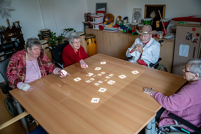 Drei ältere Frauen und ein Mann mit Hut sitzen um einen Tisch, auf den Wörter und Buchstaben projiziert werden.