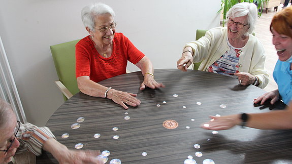 Drei Frauen sitzen lachend an einem runden Tisch, auf den bunte Bilder projiziert werden.