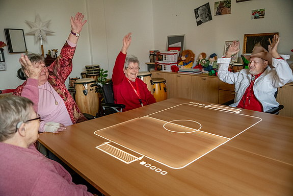 Vier ältere Menschen sitzen um einen Tisch, auf den ein Fußballspiel projiziert ist und jubeln mit erhobenen Armen.
