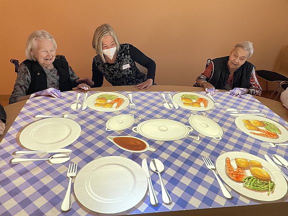 Zwei Seniorinnen und eine Frau mit Mund-Nasen-Schutz sitzen nebeneinander an einem Tisch, auf den täuschend echt Teller, Besteck und Schüsseln mit Speisen projiziert werden.